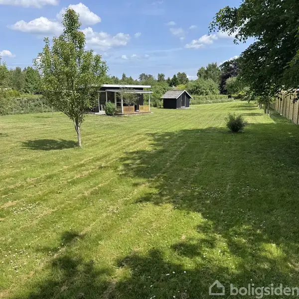 En velplejet græsplæne strækker sig i et åbent område med to træer; i baggrunden ses en lille åben pavillon og et skur under en delvis skyet himmel.