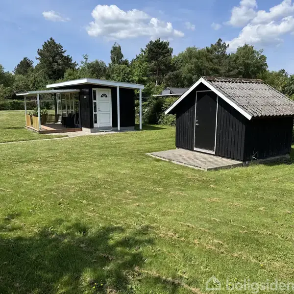 En sort, lille udhus står på en velplejet græsplæne ved siden af en mindre sort og hvid pavillon. Baggrunden består af træer og buske under en blå himmel.