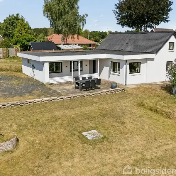 Hus står stationært, omgivet af en sparsommelig græsplæne og flisebelægning. Omkringliggende træer og buske danner et roligt boligområde. Det udendørs spisebord står på terrassen.