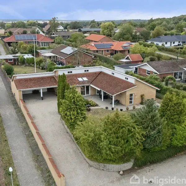 En enetageejendom med rødt tag og garage ligger på en velplejet grund med grønne buske. Husene omkring har grønne haver, og vejen er omgivet af flere beboelseshuse.