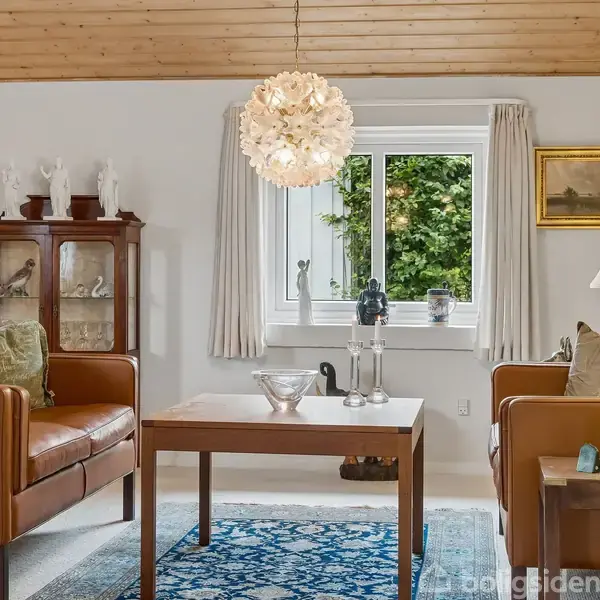 To brune lædersofaer står omkring et træbord, indrettet med lamper og dekorationer. En krystalformet lysekrone hænger fra loftet. Baggrund: hvidvæg, glasvitrine, vindue med grøn udsigt.