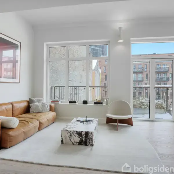 Brun lædersofa står ved vindue i lyst rum med hvid væg og moderne indretning. Lavt sofabord i marmor står på tæppe. Udgang til balkon med udsigt til bygninger.