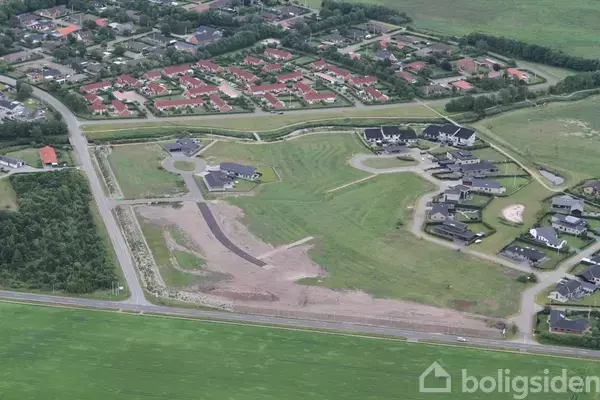 Luftfoto viser et område med nybyggeri, hvor flere huse er spredt langs kurvede veje omgivet af græsarealer, tæt på en større, etableret bebyggelse med røde tage.