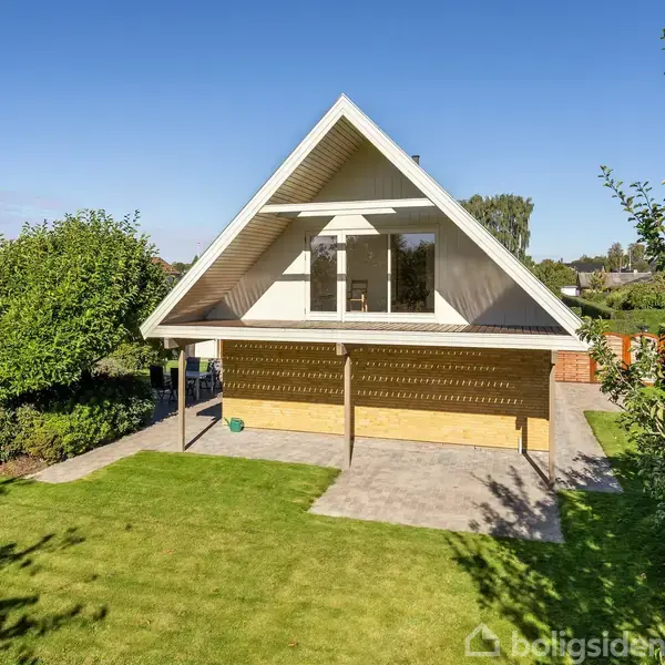 Et hvidt, trekantet hus med balkon står i en velplejet have. Balkonens skygge dækker en del af terrassen. Træer og buske omgiver huset i et roligt nabolag.