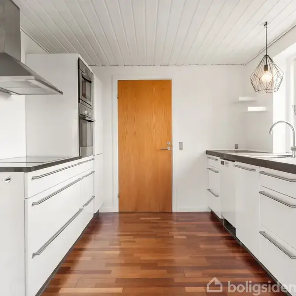 Et moderne køkken med hvidt interiør, hvor en emhætte hænger over komfuret. Dobbeltvindue lader lys ind, og en trædør fører til et andet rum. Gulvet er af mørkt træ.