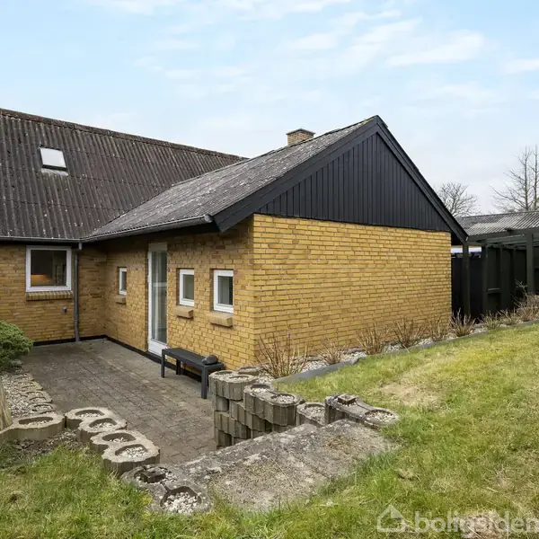 Murstenshus med sort tag og vinduer, står i en lille, delvist indhegnet have. Der er en flisesti ved siden af huset og græsplæne omkring.