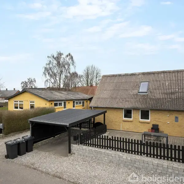 Et gult murstenshus står stille i et beboelseskvarter med en carport ved siden af, omgivet af hække og nabohuse, under en blå himmel. Vejen er foran huset.