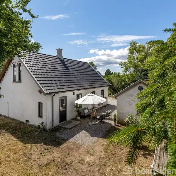 Et hvidt hus med mørkt tag står i solskin, omgivet af frodige træer. Foran huset er der en terrasse med et bord og en parasol i en haveindstilling.