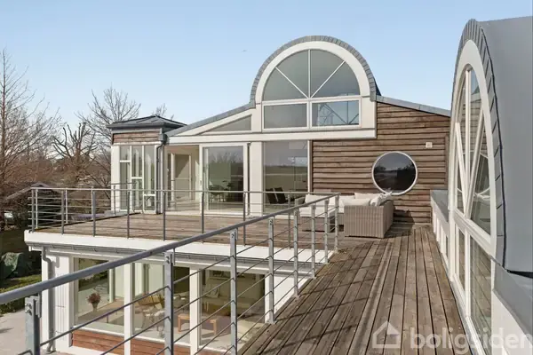 Et moderne hus med store glasvinduer og træterrasse. Vinduerne har buet design. Der er en sofa på terrassen, omgivet af træer og klart himmel.