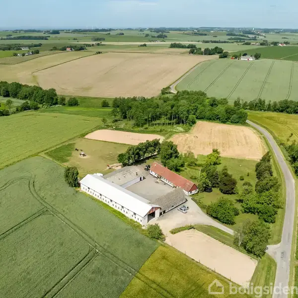 En gård med hvid bygning og rødt tag ligger omgivet af marker og træer i et landligt område. En vej snor sig forbi gården, der er placeret midt i naturen.