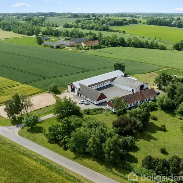 Gårdbygning set fra luften omgivet af grønne marker og træer, beliggende ved en vej i et landligt landskab under en blå himmel.