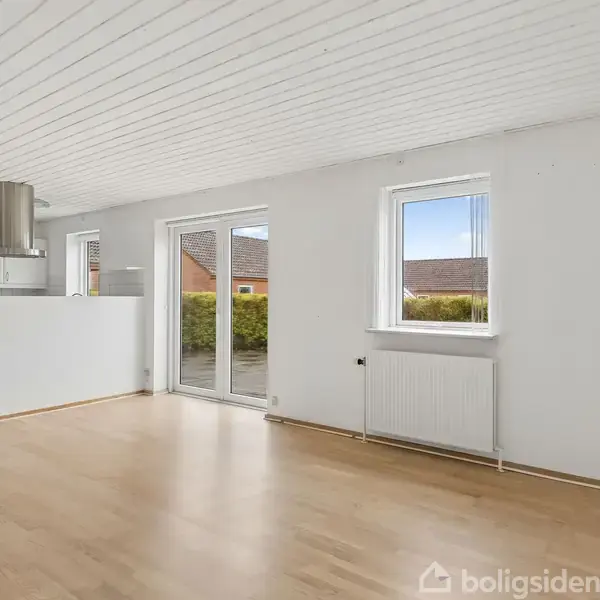 Stuen har lyse trægulve og hvidt panelloft, åbner ud mod en terrasse gennem glasdøre og har et vindue. Køkkenet er åbent med emhætte og hvide skabe.
