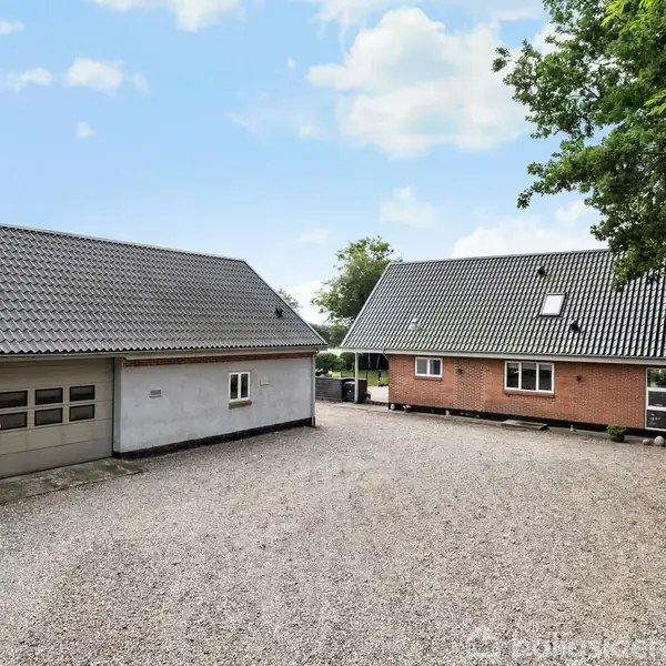 To bygninger med skråtag står ved siden af hinanden på en grusbelagt gårdsplads, omgivet af grønne træer og en blå himmel med få skyer.