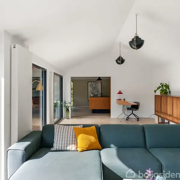 Stuen har en blå sofa med gule puder, tre hængende lamper og et skab med planter. Store vinduer giver lysindfald fra terrassen udenfor.