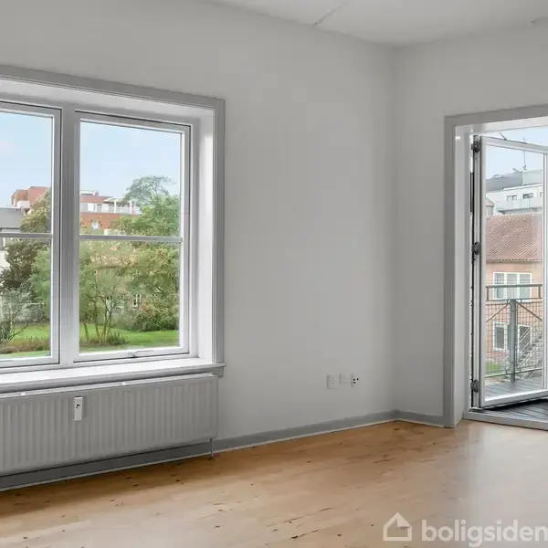 Et tomt rum med trægulve er oplyst af et vindue og en åben dør, der fører til en balkon med udsigt over en grøn have og nærliggende bygninger.