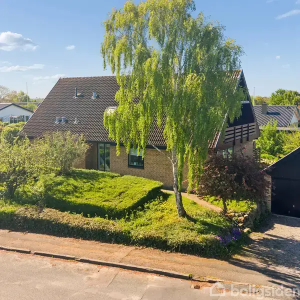 Et murstenshus med sort tag står omgivet af frodige træer og buske. En bred indkørsel fører til en sort garage. Huset ligger i et roligt boligkvarter.