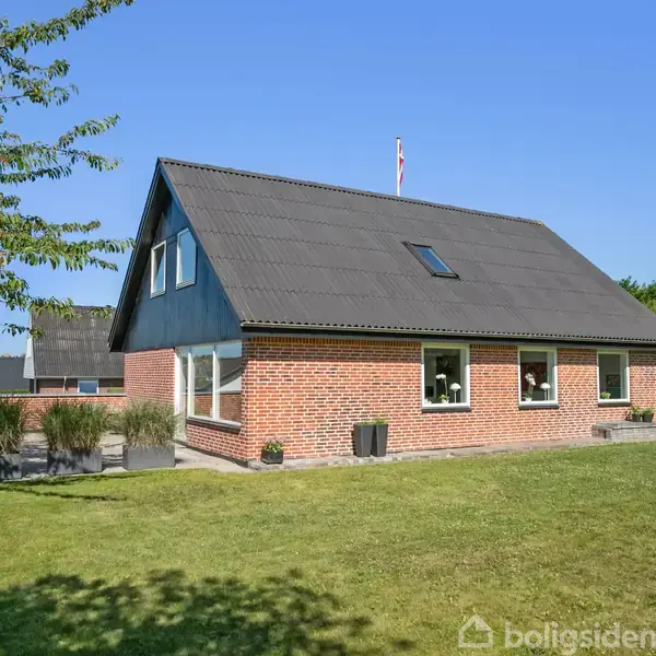 Rødt murstenshus med sort tag ligger på en grøn græsplæne. Huset har flere vinduer og et flag i baggrunden. Skyfri blå himmel og et træ til venstre.