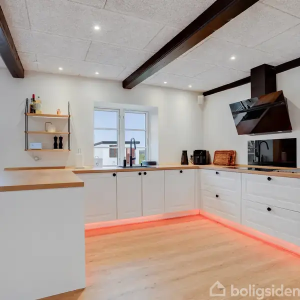 Køkken med hvidt kabinet og træbordplader. LED-lys under bordene skaber en glød. Køkkenudstyr placeret langs væggene, med sorte skabs- og skuffehåndtag. Rummet har hvide vægge og synlige loftsbjælker.