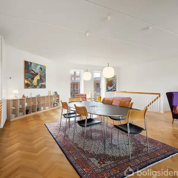 En stor spisebord står på et tæppe i en moderne stue med trægulv. Omkring bordet er der stole. Reoler med bøger og en lilla sofa ses i baggrunden.