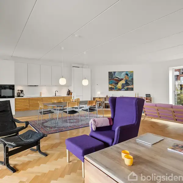 En moderne stue med en lilla lænestol og sort loungestol, hvor der står et sofabord med gule kopper. Køkken og spisebord i baggrunden. Maleri hænger på væggen.