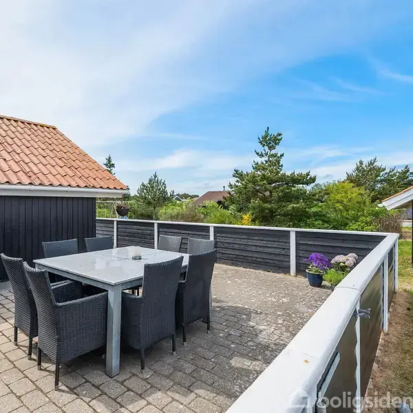 Terrassemøbler, herunder et bord og seks stole, står på en stenterrasse ved siden af et sort træhus med rødligt tag. Omgivet af grønne planter og blå himmel.