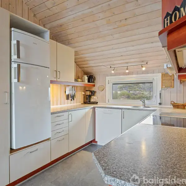 Køkken med lys indretning og skråt loft, indeholder hvide skabe, køleskab og integreret komfur. Et vindue åbner udsigt til grønt landskab, mens en hylde har teksten "HOME" på.