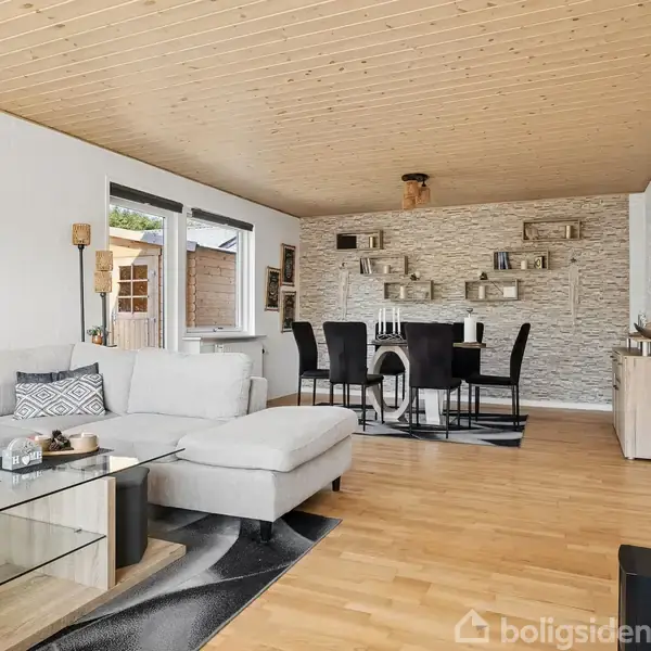 Gråt hjørnesofa står i en stue med trægulve, glas sofabord, og lyse vægge. Bagved er et spisebord med sorte stole og hylder på væggen.