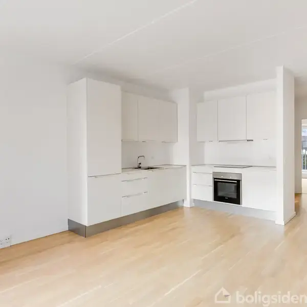 Køkken med hvide skabselementer, indbygget ovn og vask, står i et lyst rum med trægulv. Åben dør baggrund leder til en terrasse med udsigt til flere bygninger.