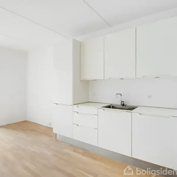 Køkken med hvide skabe og en vask i et tomt rum med lyse trægulve. Dør åbner mod en terrasse med udendørsareal dækket af fliser. Ingen tekst i billedet.