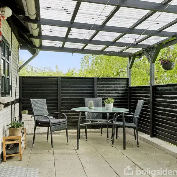 En overdækket terrasse med et rundt glasbord og tre stole. Væggene er sorte, og der hænger planter. Terrassen har flisebelægning og er omgivet af et frodigt grønt landskab.