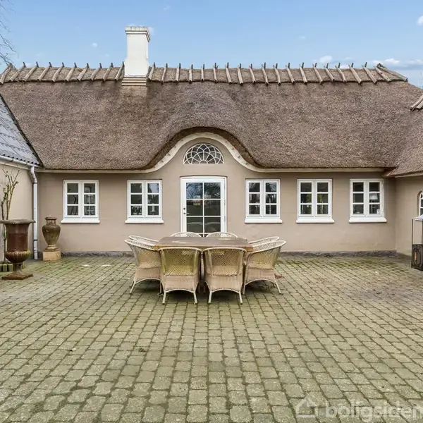 En gårdhave med et sæt møbler bestående af seks stole og et bord står på en brostensbelagt terrasse foran et hus med stråtag og hvide vinduer.