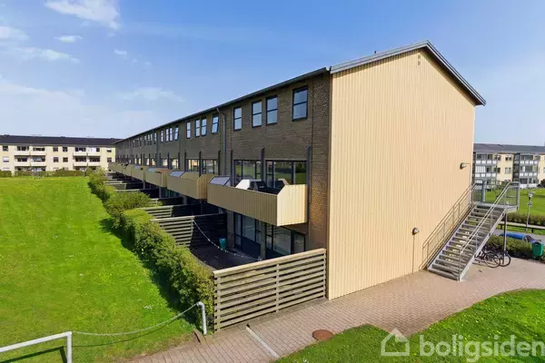 Rækkehus med flere etager og altaner; placeret i et grønt område med græsplæne og stier. Bygningen har trappeopgang og cykelparkering, omgivet af andre boliger i baggrunden.