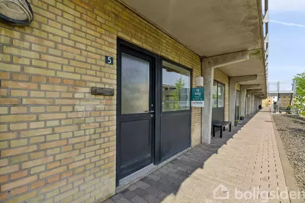 Dør med nummer 5 i sort ramme, skilt ved siden hvor der står "Til salg, Home," på gule murstenshus. Beliggende på gangbro med bænke.