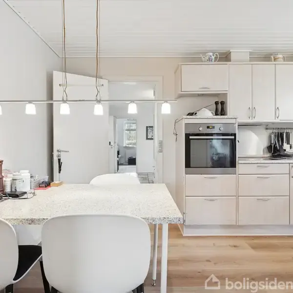 Køkken med hvidt spisebord og to stole i forgrunden, hængende lamper over bordet. Baggrunden består af hvide skabe, indbygget ovn og træoverflader på gulvet.