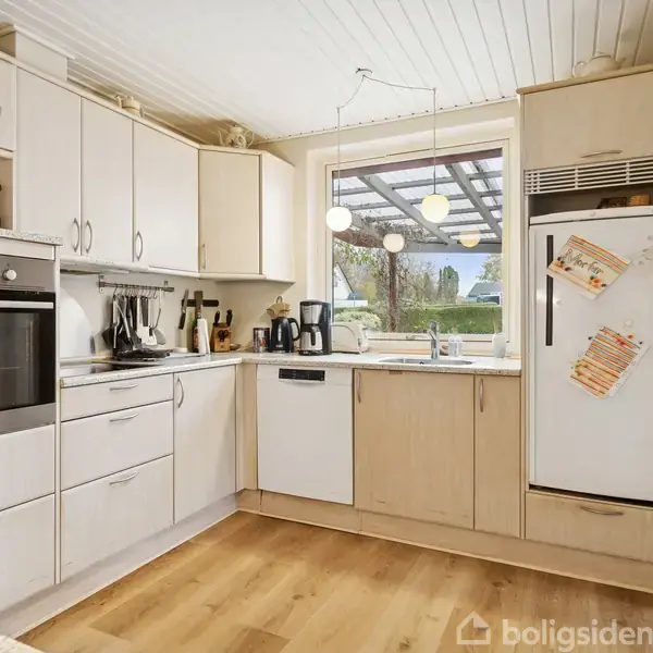 Køkken med hvide skabe og træbordplade, står i naturligt lys fra vindue. Komfur og opvaskemaskine indbygget, køleskab med magnetiske billeder. Lys trægulv giver en moderne atmosfære.