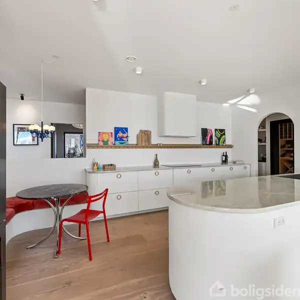 Et moderne køkken med en hvid køkkenø og indbygget vask i forgrunden. Køkkenet har indbygget skabe, en farverig vægdekoration, et rundt bord med røde stole og trægulve.