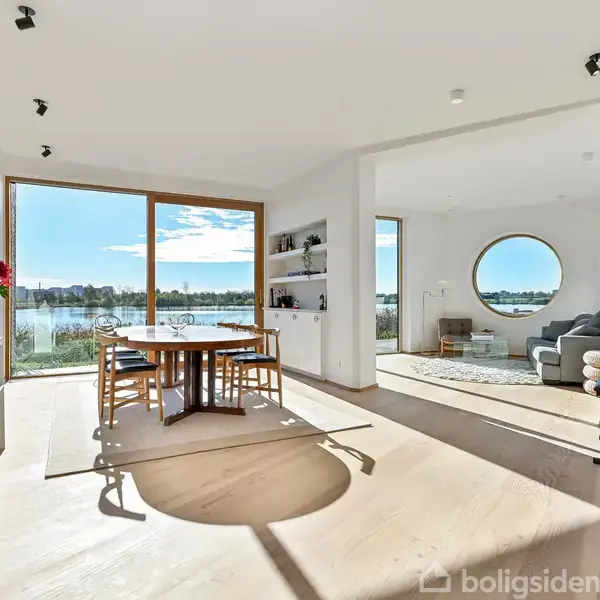 Et lyst, moderne spiseområde med et bord og stole ved store vinduer, der viser udsigt over en sø. Til højre er en stue med rundt vindue og sofa.