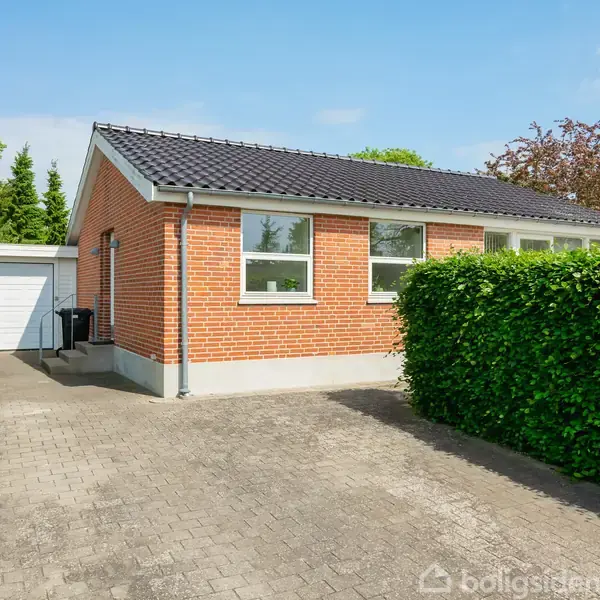 Rødt murstenshus med sort tag står stille ved siden af en hæk. En flisebelagt indkørsel fører op til en hvid garage. Lyst og grønt haveområde omgiver huset.