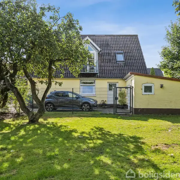 Et gult hus med mørkt tag står i en have med grønne træer. En bil er parkeret foran huset, omgivet af græsplæne og et hegn.