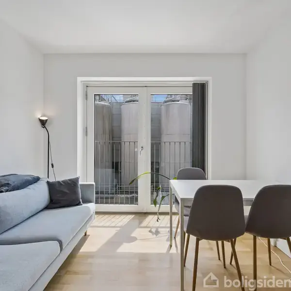 Sofa i lyseblå farve står til venstre, ved siden af et lille spisebord med grå stole. Rummet er lyst med parketgulv, og glasdøre åbner til en lille altan.