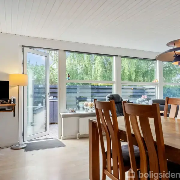 Spisebord omgivet af træstole står i et lyst rum. Dør til venstre åbner ud mod en terrasse. En bordlampe lyser op ved siden af et vindue med udsigt til have.