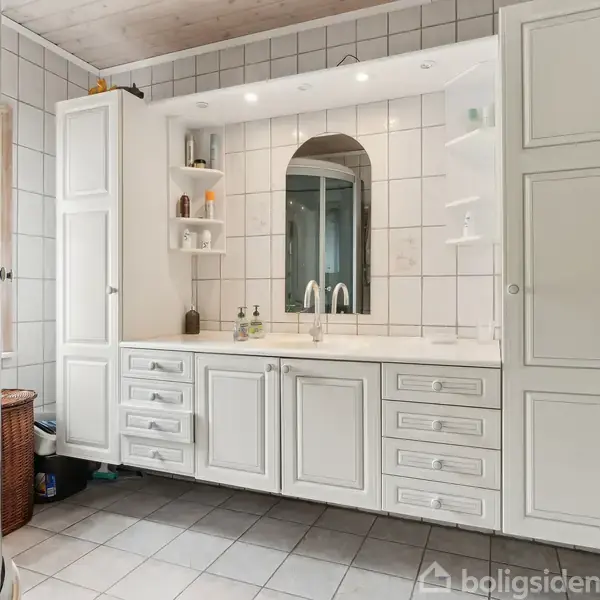 Hus med hvid facade og rødt tag står langs en stille gade, omgivet af lavtliggende bygninger. En carport ses ved venstre side, mens en blå himmel danner baggrunden.