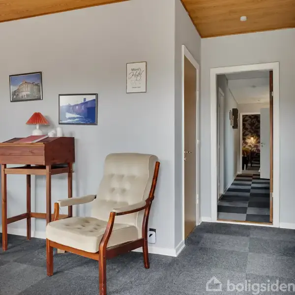To lænestole står ved en væg med indrammede billeder, mens en lamineret gangsti fører til en anden del af huset. En lille træreol med en lampe står ved siden af stolene.