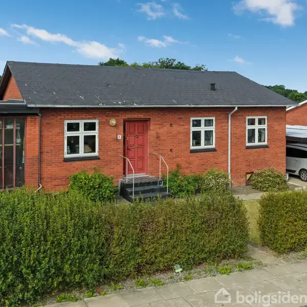 Rødt murstenshus med sort tag, har rødt indgangsparti og tre vinduer. Foran er en velplejet hæk langs fortovet. En carport med en bil er til højre.
