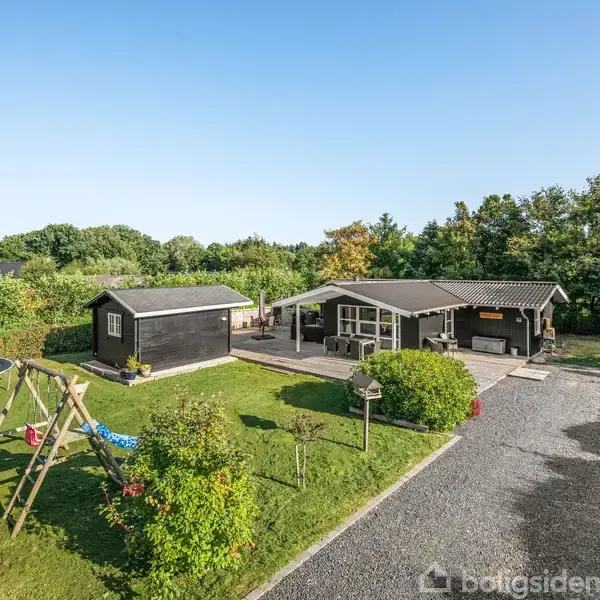 Et sortmalet sommerhus med terrasseluft sidder omgivet af grønne buske og træer. En legeplads med gynger og en trampolin står til venstre. Grusvej fører til huset.