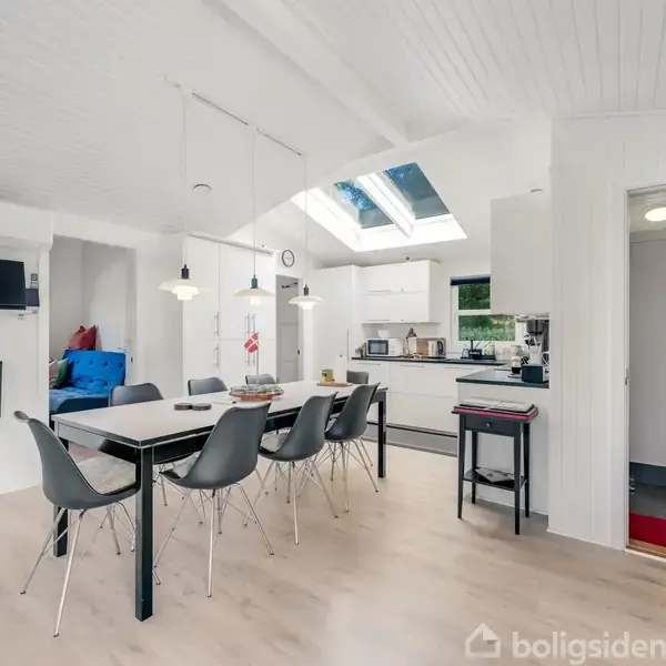 Spisebord med seks stole står i et lyst, åbent køkkenalrum med hvide vægge og loft. Et ovenlysvindue i loftet slipper naturligt lys ind. Der ses også et åbent badeværelse.