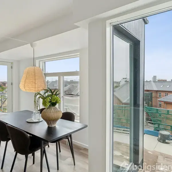 Spisebord med fire stole står i en lys spisestue. Bordet er pyntet med en vase med planter. Store vinduer giver udsigt til en by med røde teglstenstag.