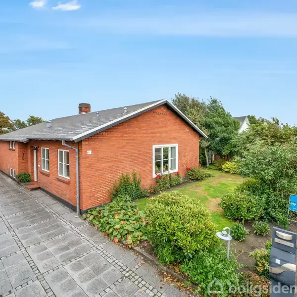 En rød murstensbungalow står stille ved en flisebelagt indkørsel. Omgivet af en velplejet have, der grænser op til en vej.