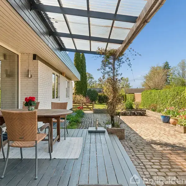 En overdækket terrasse med et træbord og fire stole står ved siden af en hvid murstensbygning. Baggrunden består af en velplejet have med stier og blomsterbede.