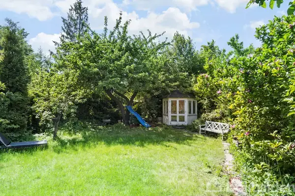 En have med grøn græsplæne og lyseblåt legehus i træ; omgivet af tætte træer og buske. En hvid bænk og rutsjebane ses ved siden af legehuset i baggrunden.